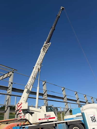 A mobile crane is positioned at a construction site in Wilkowisko, Poland, lifting steel beams for a new building. The crane's white boom is extended upwards against a clear blue sky. The partially constructed building features a steel framework of vertical beams and horizontal supports. In the foreground, the crane's cab is visible, with company logos and a phone number displayed. Adjacent to the crane is a portable toilet unit. The ground is a mix of earth and grass, with trees in the background. The scene suggests ongoing construction activity under fair weather conditions.