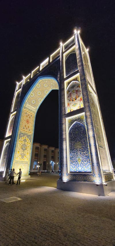 This is a night-time, low-angle shot of an ornate archway decorated with intricate carpet-like patterns and illuminated by a string of lights. The archway features geometric designs and floral motifs in shades of blue, yellow, and red, set against a dark background. The ground beneath the archway is paved with cobblestones. Several people are visible in the lower right portion of the frame, standing near the base of the archway and seemingly interacting with each other. Their presence suggests the archway is accessible and perhaps a point of interest or gathering. The overall impression is one of grandeur and cultural artistry, illuminated against the night sky, with the surrounding buildings in the background appearing less elaborate. The scene appears calm and peaceful. There is no visible text in the image.