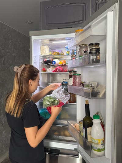 In a kitchen in Novoaleksandrovka, Russia, a young woman with blonde-streaked brown hair, wearing a black t-shirt, is standing in front of an open refrigerator. She is holding a clear plastic bag filled with green leafy vegetables, and her right hand is reaching into the refrigerator. The refrigerator is well-stocked with various food items, including jars of condiments, bottles of drinks, eggs, and other produce. The kitchen has gray walls and cabinets, with a light fixture visible on the ceiling.