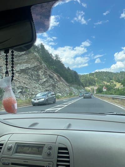 The image shows a view from inside a car driving on a road. The road is asphalt with white lane markings. There are several cars on the road ahead. On the left side of the road is a steep, rocky embankment covered with vegetation. On the right side of the road is a metal guardrail and then a forested hillside. The sky is blue with scattered white clouds. In the foreground, the dashboard and radio of the car are visible. A car air freshener hangs from the rearview mirror. The scene appears to be a highway or a main road through a mountainous or hilly area, consistent with parts of Balabanovo, Bulgaria.