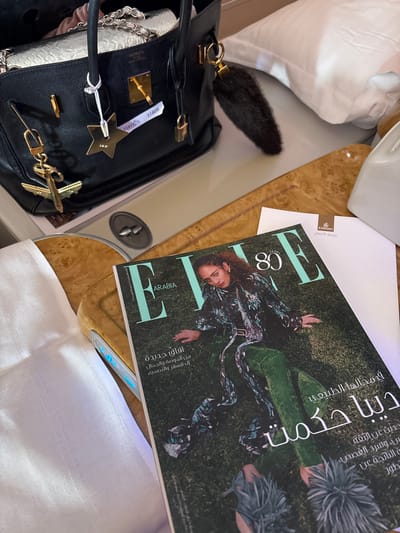 The scene is set within the business class cabin of an Emirates aircraft, with the broader contextual location specified as the city of Abqaiq, Saudi Arabia. A black leather Hermès Birkin handbag is positioned on a light wood-grain veneer surface, which appears to be a deployed aircraft tray table. The handbag features prominent gold-colored hardware, including the Hermès stamp, a lock, and a key clochette. It is accessorized with a gold-colored metal charm depicting an airplane attached to an anchor, a gold-colored star-shaped charm with a small white tag displaying "31808," and a dark brown fur-like bag charm on its right handle. Partially visible inside the bag are light-colored items, including a silver metallic object and pink fabric. To the right of the handbag, an "ELLE Arabia" magazine lies face-up on the tray table, featuring a woman with dark curly hair on its cover, dressed in a patterned coat and green velvet pants, identified by Arabic text as "Dina Hikmat." A white paper or menu, bearing the Emirates logo and "Business Class" in Arabic ("درجة الأعمال"), is partially visible beneath the magazine. White pillows and a white seat covering are visible in the background. A portion of a white linen napkin or blanket covers the left armrest, which displays blue illuminated control buttons. No individuals are present in the image.