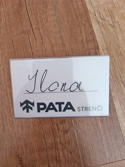 A rectangular name tag is placed on a wooden surface. The name tag has a white background. Handwritten in black cursive script on the top half of the name tag is the name "Ilona". Below the name, a horizontal line is drawn. The bottom half of the name tag features a logo consisting of a stylized green tree silhouette to the left, followed by the word "PATA" in large black capital letters. To the right of "PATA", the word "STRENČI" is written in smaller, light gray capital letters. The wooden surface has a natural grain pattern and appears to be a floor or tabletop. The lighting suggests an indoor setting. The name tag is likely a personal identification or designation tag, possibly used at an event or workplace. The location of Tebras, Latvia is not discernible from the image itself.