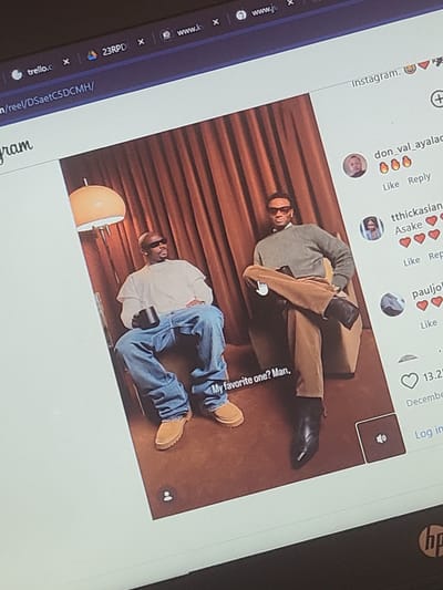 The image is a photograph of a laptop screen displaying an Instagram reel featuring two men posing indoors. The screen, part of an HP laptop, shows a web browser interface with multiple tabs open, including "trello.c", "www.k", "www.ju", and a URL indicating an Instagram reel ("/reel/DSaetC5DCMH/").
The Instagram reel shows two men lounging against a dark red, pleated curtain-like backdrop. The man at the top is lying on his side, wearing a grey long-sleeved top, tan trousers, and black boots, with what appear to be glasses. The man at the bottom is lying on his back, dressed in a white t-shirt, blue jeans, and tan shoes, wearing sunglasses and holding a dark device. A lamp with an orange dome shade is visible near the second man. Both appear to be posing in a relaxed manner.
Visible text on the Instagram interface includes "Instagram.", usernames like "don_val_ayalad", "tthickasian", and "pauljol", alongside options such as "Like Reply" and the word "Asake". A caption or comment snippet reads "My favorite one? Man,", with a like count of "13.2" and "Decembe" also visible. A hand-shaped cursor is present on the screen, pointing at the upper man's chest area. Other visible text includes "23RPD", "ram", and "Log in".
Location context provided: Oke Ogun, Nigeria.