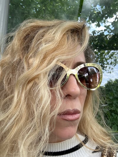 A woman with blonde hair is lying on a surface, likely indoors, with a window showing trees and sky. She is wearing sunglasses with gold frames. Part of a black and white striped shirt or sweater is visible. The location is indeterminable in the city of Dronten, Netherlands.