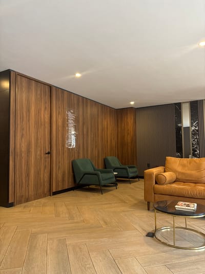 An interior space features a tan sectional sofa with a round glass-topped table beside it. Two green chairs with black frames are positioned near a wood-paneled wall, which also includes a list of text behind glass. The floor transitions from wood paneling to herringbone-patterned planks, and a white wall extends vertically with recessed lighting. A dark border edges the base of the wood paneling.