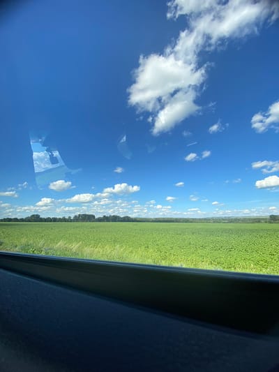 The image depicts a vast, verdant field of crops under a clear blue sky dotted with fluffy white clouds. The field appears to be agricultural, with uniform rows of green plants stretching towards a distant treeline. The foreground is dominated by the dark, curved surface of a vehicle's interior, likely a dashboard or door panel, suggesting the photo was taken from inside a moving vehicle. Reflections of the sky and clouds are visible on the windshield or window. The overall impression is one of a scenic, rural landscape. Based on provided metadata, this scene is located in Isyangulovo, Russia.