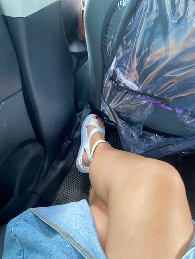 A person's legs are visible inside a vehicle. The person is wearing white sandals and a denim skirt. The interior of the car, including the dark grey plastic paneling and a seat covered in clear plastic, is visible. The floor of the vehicle has a textured mat. The person's right leg is bent at the knee and resting on the left leg, with the foot pointing towards the right. The lighting suggests it is daytime. There is no visible indication of the location being Yuldybayevo, Russia.