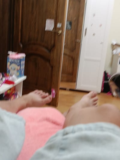 The image depicts an indoor scene with a focus on a person's legs and feet, positioned on a pink surface. In the background, there are wooden doors, one of which appears to be slightly ajar, and a white closet door with some colorful stickers. To the left, a white shelf holds a blue box with a cartoon character and other items. The lighting suggests an indoor setting, possibly a bedroom. The image does not provide any specific information to determine its location in Novotitarovskaya, Russia.