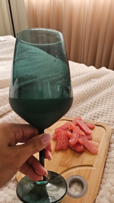 A hand with pink-painted fingernails holds the stem of a dark green wine glass filled with a dark liquid. To the right of the glass, a small wooden cutting board displays a pile of pink, rectangular candies. The scene is set on a textured white blanket, with soft, draped curtains in the background. The image appears to be taken indoors, likely in a residential setting. There is no information available regarding the location in the city of Slengiai, Lithuania.