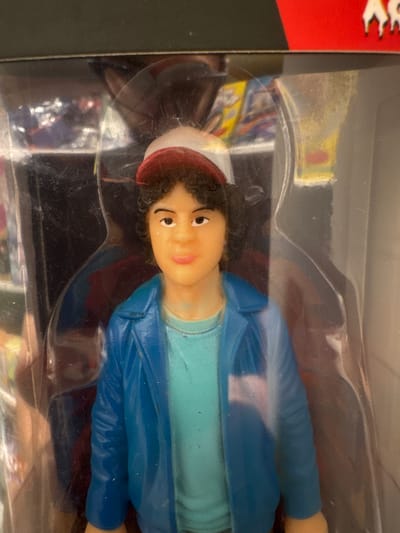 This media file shows a close-up, vertically oriented shot of a toy figure of a young boy, likely Dustin Henderson from the TV show "Stranger Things," encased in clear plastic packaging. The figure is positioned horizontally, with its head and upper body most prominent. The toy has curly brown hair, a light skin tone, brown eyes, and is wearing a light blue t-shirt under a royal blue jacket. A red and white baseball cap is on its head, tilted to the side. The background within the packaging is blurry, but suggests other toys or merchandise, possibly with colorful illustrations. The exterior packaging is clear plastic with some smudges and dust visible. The left side of the frame shows a sliver of red packaging with white jagged designs. The overall impression is that of a new toy being presented for sale. There is no visible text in the immediate frame. The location context provided is Keila, Estonia, which is not visually discernible from the image itself. The lighting appears to be artificial, and the time of day and weather are not determinable. The emotion conveyed by the toy's facial expression is neutral.