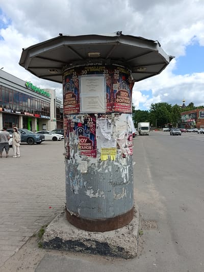 The image shows a cylindrical advertising pillar covered in torn posters and graffiti, situated on a sidewalk next to a paved road in Koltushi, Russia. The pillar is topped with a weathered, octagonal metal canopy. To the left of the pillar is a brick building with large windows, featuring a green "Perekrestok" supermarket logo and other commercial signs. Several cars are parked on the sidewalk. Two individuals, a man and a woman, are standing and facing the building. The road in the background is asphalt and appears to be a moderately busy street with a white truck and several other vehicles. Trees and buildings, including a church with a golden dome, are visible in the distance. The sky is partly cloudy with a mix of white clouds and blue sky.