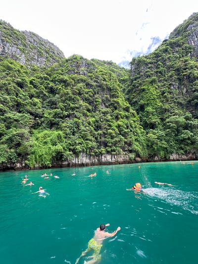 The image shows a group of people swimming and snorkeling in a body of turquoise water, with steep, lush green cliffs rising behind them. The cliffs are covered in dense vegetation, with some exposed rock faces visible. The sky is overcast with some patches of blue and white clouds. The water is calm and clear, with ripples visible from the swimmers. Several people are visible in the water, some with snorkeling masks and fins, others without. One person in the foreground is wearing a yellow and green patterned swimsuit, another is wearing a life jacket. In the background, the cliffs form a dramatic backdrop to the scene, with the vegetation creating a rich green texture. The overall impression is one of a tropical paradise. This scene is likely located in Haad Yao, Thailand, a popular tourist destination known for its beautiful beaches and clear waters.