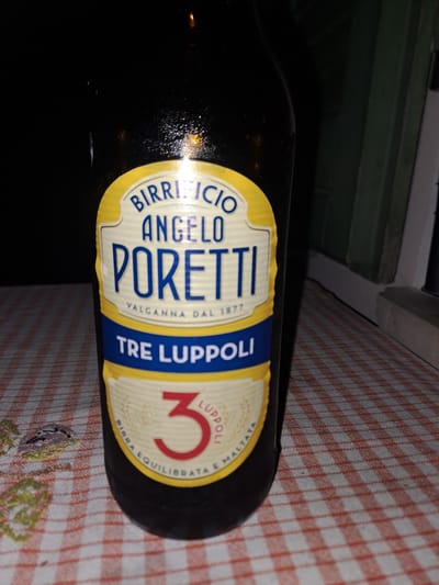 A close-up, slightly angled, eye-level shot showcases a dark glass beer bottle with a prominent label. The label is yellow and blue with white text. At the top of the label, in blue text, are the words "BIRRICICIO ANGELO". Below this, in larger blue text, is "PORETTI". Beneath "PORETTI", in smaller white text, is "VALGANNA DAL 1877". Further down, in a blue band, reads "TRE LUPPOLI". Below this, a yellow band surrounds a red numeral "3". Encircling the numeral "3" is text in a light beige color, reading "BIRRA EQUILIBRATA E MALTATA". The bottle is placed on a table covered with a red and white checkered tablecloth. The background is dark and out of focus, with faint suggestions of a window frame on the right. The scene does not depict any people or specific city locations.