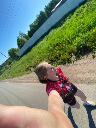 A person is running on a paved path, likely a road or trail. The runner is wearing a red and black running shirt and black shorts. They are wearing running shoes with yellow accents. The runner appears to be holding a phone or camera, taking a selfie while running. The location is rural, with green grass and a long wall visible on the left. The sky is blue. It is not possible to identify the location as Kukui, Russia, from the visual information.