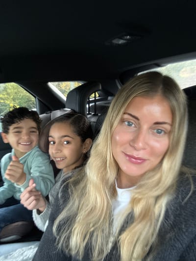 The image captures three individuals inside a vehicle, identified as being in Pourrières, France. In the foreground, an adult female with long blonde hair and blue eyes, wearing a white shirt under a dark gray cardigan, looks towards the camera with a slight smile, suggesting she is taking a selfie. In the back seat, two children are visible. On the left, a boy with dark curly hair, wearing a light blue-green long-sleeved top, smiles broadly and gives a thumbs-up gesture with his left hand. Next to him, a girl with dark hair, wearing a light gray sweater, smiles and forms a finger heart gesture with her right hand. Both children appear seated in the rear, looking at the camera. Through the car's rear window, blurred green and yellow foliage is visible, indicating an outdoor setting. The car interior features dark upholstery and headrests.