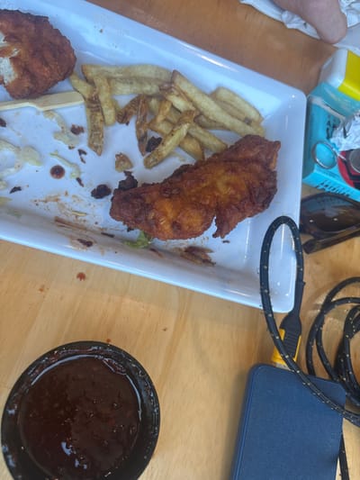 A mostly empty rectangular white plate contains remnants of a meal: french fries, fried chicken pieces, and shredded lettuce. A black bowl filled with a dark, viscous sauce sits to the right of the plate on a wooden table. A charging cable and a blue electronic device are visible in the upper right corner. Sunglasses and other items are scattered to the left and top of the plate. The overall setting suggests a casual dining experience.