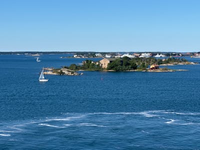 The image depicts a bright, sunny day with a clear blue sky over a body of water. In the foreground, the deep blue water exhibits some white foam trails, suggesting movement.
In the middle ground, a small, rocky island is visible, densely covered with green trees. On this island, there is a rustic stone building, appearing to be a ruin, and a small, orange-roofed building with red walls. Further to the right, another small island features a single red-roofed house.
Beyond the islands, the water extends to a distant coastline. On this coastline, various buildings and structures are visible, including what appear to be docks and several large ships, some of which have white hulls and superstructure. The scene suggests a port or naval base area.
Two sailboats are present in the water. One is closer to the foreground, a white sailboat with its sail up, moving from left to right. Another smaller white sailboat is visible further in the distance, near the islands. A red buoy is also present in the water.
The overall impression is of a picturesque coastal landscape with a mix of natural beauty and human activity, likely related to maritime operations. The location is identified as the southern archipelago of Karlskrona, Sweden, a UNESCO World Heritage site known for its naval history and picturesque islands.
