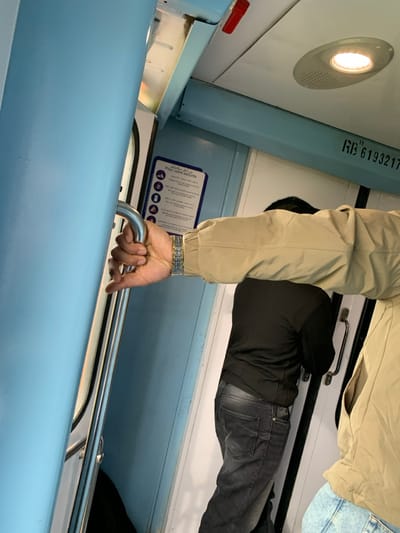 The image displays the interior of a train carriage, consistent with public transportation within Sidi Kacem, Morocco. The environment features light blue walls and a white ceiling equipped with a circular illuminated fixture and a red emergency handle. A numeric identifier, "RB 6193217," is stenciled on the upper wall/ceiling interface.

In the foreground, the right hand of an individual is gripping a silver-colored vertical grab rail. This person wears a silver-toned metal link watch on their left wrist, a light tan or beige jacket, and light blue jeans, partially visible.

Further inside the carriage, a second individual is observed from the rear, wearing a black long-sleeved top or jacket and dark grey jeans. This person is standing near a door equipped with a horizontal handle. A rectangular sign on the wall displays safety pictograms and text in both Arabic and French, including "Pour votre sécurité." No direct interactions between the individuals are depicted.