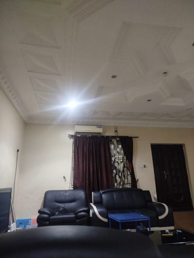 The image depicts an interior scene of a living room, likely located in Sapele, Nigeria. The room features a white, intricately designed ceiling with recessed square patterns and decorative molding along the edges. A bright light source, possibly an overhead lamp, is visible in the upper portion of the image, causing glare.
Furnishings include two black leather sofas. The sofa on the left is partially visible and appears to be a single-seater. The sofa on the right is a two-seater with white accents along its arms. In front of the right sofa sits a small, bright blue rectangular coffee table. Next to it, a black glass-topped coffee table is partially visible, with some items on its surface. Behind the sofas, a dark brown curtain is drawn across a window, and an air conditioning unit is mounted on the wall above it. To the right of the curtained window is a dark brown door. On the left side of the room, a portion of a television screen and a thin black pole are visible, suggesting a media setup. Other items on the floor include plastic bins and what appears to be a dark bag under the blue coffee table. The walls are painted a light, neutral color. There are no people visible in the frame.