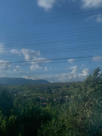 The image displays a panoramic view of a rural mountainous landscape in Yasinia, Ukraine, under a clear blue sky populated with scattered white cumulus clouds. Multiple dark, thin horizontal wires, consistent with utility lines, traverse the upper portion of the frame, partially overlaying the sky and distant hills. The immediate foreground is dominated by dense, dark green foliage from trees and bushes, which partially obscures the lower midground.

The midground reveals a settlement or village nestled within a valley and across rolling green hills. This area contains numerous residential structures with dark roofs, interspersed with smaller trees and patches of green grass. Further into the midground, the hills rise, covered in green vegetation, with some lighter patches indicating open ground.

In the background, larger, darker mountains with varied terrain are visible, their peaks partially obscured by the sky or distant haze. On a prominent hill in the mid-distance to the right, two slender, dark vertical structures, consistent with communication towers or electrical transmission pylons, are present. A large, light-colored, dome-shaped or building structure is also visible on a more central distant hill. No human figures, animals, or vehicles are discernible in the scene.