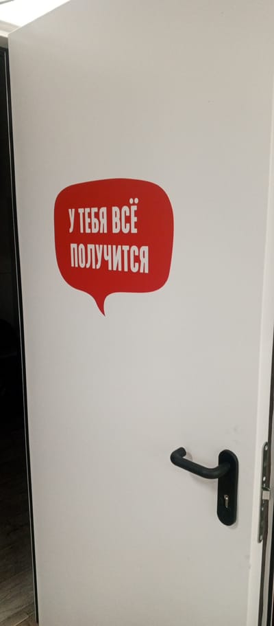 A white door with a red speech bubble sticker is shown. The sticker contains white text in Russian that reads "У ТЕБЯ ВСЁ ПОЛУЧИТСЯ" which translates to "You will succeed." A black door handle is visible on the right side of the door. The background is a plain white surface. The image is an indoor shot, likely in a residential or office setting. There are no people or animals present in the image.