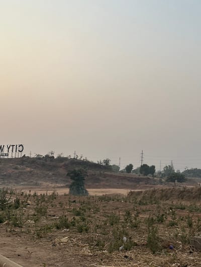 Hazy landscape featuring a dirt embankment covered in dry vegetation. Utility poles stretch into the distance. A dirt path runs along the base of the embankment. The sky is overcast, appearing pale and washed out. A sign, partially visible, reads "CITY W..." near the bottom left corner. The overall scene evokes a sense of dry, dusty environment.