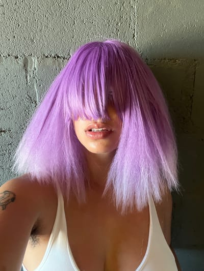 The image presents a close-up, chest-up portrait of an individual, likely female, situated in Arraial Do Cabo, Brazil. The subject is characterized by voluminous, bright purple hair with full bangs that completely obscure the eyes and upper facial features. The hair transitions subtly to a lighter lilac or white at the ends. The visible lower face displays full, glossy lips, slightly parted to reveal upper teeth fitted with dental braces. The individual wears a white tank top, with a thin strap visible over the right shoulder. Dark, visible hair is present in the right armpit region. A small, dark-outlined tattoo, appearing abstract or avian, is visible on the upper right arm/shoulder. The background is a textured, grey concrete or cinder block wall, with lighting suggesting illumination from the viewer's left, creating a darker area on the right side of the background.