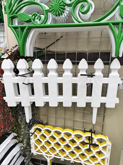 This image depicts a retail display of garden decorations. In the foreground, a white plastic picket fence section is prominently displayed. Behind it, a section of yellow and white lattice-like decorative fencing is visible. Above the picket fence, a green, ornate garden decoration, possibly part of a larger piece, is hanging, featuring leaf and swirl motifs. The items are arranged on a wire grid display, common in retail environments. To the left and behind the picket fence, artificial greenery with red and purple accents is also part of the display. The background is a plain beige wall. No people are visible in the image. There is no indication or information within the image to suggest this scene is located in Mayma, Russia.