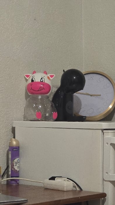 The image depicts an indoor scene within the city of Deventer, Netherlands. A light-colored, textured wall serves as the background for objects situated on two distinct surfaces.
On an elevated white surface, from left to right, a clear plastic container shaped like a stylized cow is present; it features a white head with pink ears, snout, and tongue, and pink details on its lower body. Adjacent to it, a small, black, glossy appliance or decorative figurine with a rounded, curved profile stands. To the right of this black object, a circular wall clock with a gold-colored frame and a white face displays two light-colored hands, indicating approximately 12:40.
On a lower, wooden-toned surface in the foreground, from left to right, a purple aerosol can labeled "Glade" and "J. Johnson" stands upright. Next to it, a white power strip (extension block) is horizontally positioned, with a black electrical cable plugged into one of its outlets. The silver edge of an electronic device, possibly a laptop or tablet, is partially visible on the far-left foreground of this lower surface. No individuals, explicit actions, or interactions are depicted within the static scene.