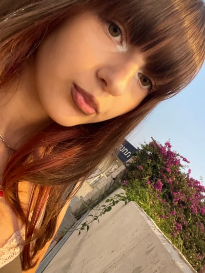 The image displays a slightly tilted selfie of a female individual captured outdoors in Kefar Sava, Israel. The subject, positioned in the upper left and center of the frame, has long brown hair with reddish-auburn highlights and bangs that fall across her forehead. Her eyes are brown, and her lips are slightly parted, revealing the tip of her tongue. She is wearing a light-colored, possibly lace or crocheted, top with a thin strap visible on her left shoulder, and a silver-colored chain necklace around her neck. Her face is illuminated by warm, golden-hour light from the right. The background, out of focus, features a light-colored concrete wall or structure in the lower right. Behind this, a dense green bush heavily adorned with vibrant pink-magenta flowers, consistent with bougainvillea, is visible. Above the foliage, a clear, pale blue sky stretches across the upper right. A dark blue sign with white or light-colored Hebrew text is partially visible in the middle right background, showing the characters "חתנן," suggesting a business or establishment within Kefar Sava.