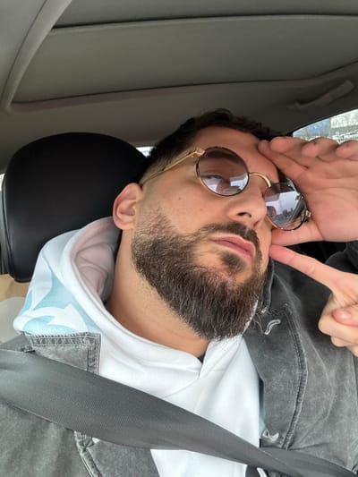 An adult male is depicted seated in the passenger seat of a vehicle. He has short, dark hair and a full, dark beard. He is wearing round, gold-framed sunglasses with reflective, dark lenses. His attire includes a white hooded sweatshirt with a light blue patterned interior visible in the hood, overlaid by a dark gray, washed denim-style jacket. A dark gray seatbelt is secured across his chest and left shoulder. The man's right hand is raised, with fingers touching his forehead above his sunglasses. Two fingers of his left hand are visible, touching his left cheek. The vehicle's interior features a black headrest and a light-colored ceiling. Through the vehicle's window, a blurred outdoor background shows indistinct foliage and structures, indicating a daytime setting with diffuse light. The image contains no discernible visual elements to specifically identify the location as Vendenheim, France.
