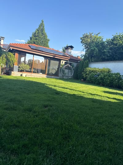 The image depicts a spacious, well-maintained garden in Malo Buchino, Bulgaria, on a clear day with a bright blue sky. A modern house with a red-tiled roof and large glass windows is visible, featuring solar panels on the roof. A swing chair hangs in the garden, adding a touch of leisure. The lawn is lush green, with shadows cast by unseen trees, suggesting it is late afternoon. A white wall encloses part of the property, and mature trees and shrubs provide privacy and natural beauty. The overall impression is one of a serene and comfortable residential setting.