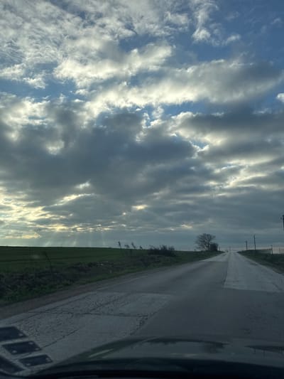 The image shows a rural road in Sofronievo, Bulgaria. The road stretches into the distance, flanked by green fields and sparse vegetation on one side, and the vehicle's interior on the other. Above, the sky is filled with dramatic, striated clouds, with glimpses of blue sky visible. The lighting suggests it is either sunrise or sunset. There are utility poles along the side of the road. The road surface appears to be wet, with some pooling of water. No people are visible.