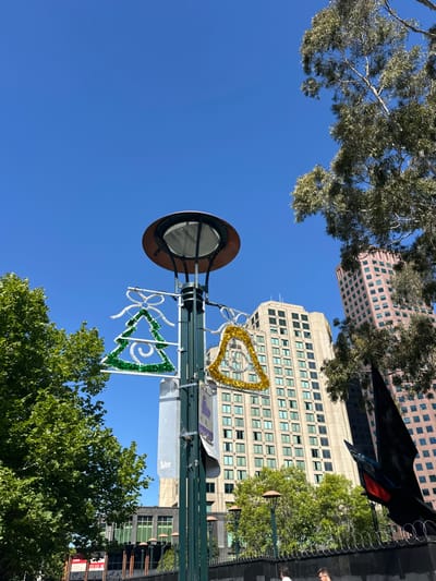 A low-angle shot captures a city scene with a tall lamppost taking center stage. The lamppost is adorned with festive decorations, including a gold tinsel triangle and a green illuminated tree shape. Buildings of varied heights surround the lamppost. The sky is bright blue, with trees visible both behind and above the lamppost, adding depth to the composition.