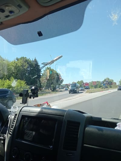 The image is taken from inside a vehicle looking out through the windshield. In the foreground, the dashboard of the vehicle is visible, including a dark screen, air vents, and a black phone mount. Outside the vehicle, a road is visible with several cars and a black van driving on it. There are trees and buildings in the background. The sky is clear and blue. A prominent object in the sky is a silver airplane on a metal pole, appearing to be a monument or art installation. This airplane sculpture is located near the city of Dubno, Ukraine. The overall scene depicts a sunny day on a highway, with the airplane sculpture serving as a distinctive landmark.