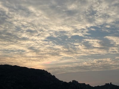 The image displays a wide-angle landscape captured in Sanapur, India, depicting a sky filled with patterned clouds over a silhouetted geological formation. The upper two-thirds of the frame are occupied by a cloud-filled sky, primarily featuring stratocumulus or altocumulus formations arranged in distinct, rippled patterns across a light blue-grey background. Sunlight, likely from a low sun angle, illuminates these clouds from below, imparting a warm, yellowish-white luminescence to their undersides, transitioning to cooler grey-blue tones higher in the sky. A subtle patch of pink and light red iridescence is visible low on the horizon, slightly left of the image center, embedded within the illuminated cloud layer. The lower third of the image shows a dark, undulating silhouette of rocky hills or mountainous terrain. The rocks appear rugged and irregular, forming a jagged horizon line against the sky. No individuals, animals, man-made structures, or direct actions are visible within the frame. The environment is natural and appears to be in an early morning or late evening phase based on the sky's illumination.