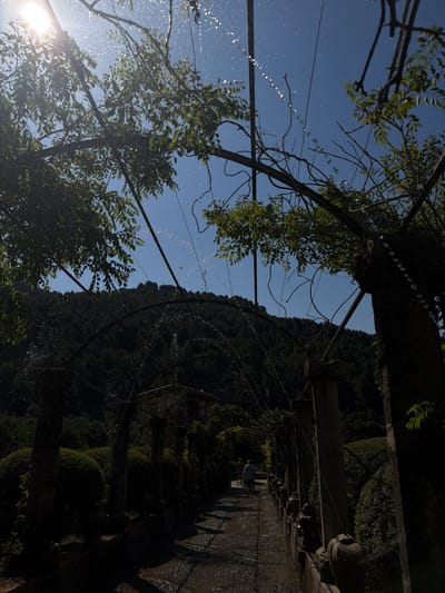 The image shows a water feature with a series of arches creating a tunnel of water droplets. The scene is partially obscured by trees, with sunlight shining through the leaves in the lower left. A blue sky is visible through gaps in the foliage and water arches. In the background on the right, a staircase or path lined with bushes and walls can be seen. At the top of the stairs, two people can be vaguely made out. The location is the Alfàbia Gardens in Bunyola, Spain.