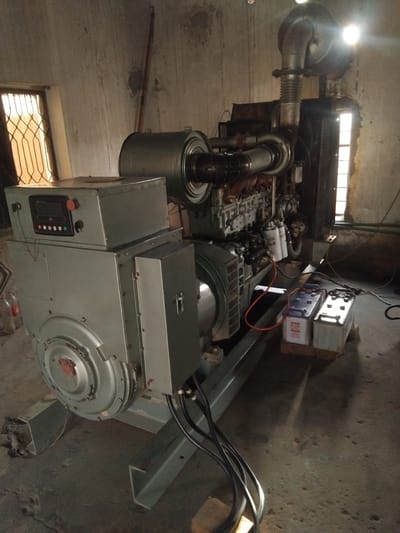 The image shows a large diesel generator set situated indoors in what appears to be a utilitarian building. The generator is gray and has a complex arrangement of pipes, filters, and a large exhaust system. A control panel with a digital display and buttons is visible on the front of the generator. Two batteries are connected to the generator with red and black cables. The environment is dimly lit with light streaming in from two windows with decorative metal grates. The walls are rough and unpainted, and the floor is concrete. There are no people visible in the image. The generator is likely located in Chhina Uttar, Pakistan, as indicated by the prompt.