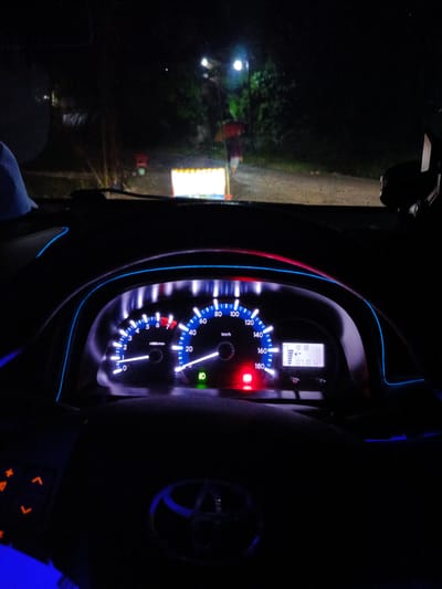 The image is taken from inside a car at night, looking out through the windshield and at the car's dashboard. The dashboard features a speedometer, tachometer, and a digital display. The speedometer reads up to 180 km/h, with the needle pointing to approximately 10 km/h. The tachometer shows readings up to 7 x 1000 r/min, with the needle near 0. The digital display shows the time as 8:18 and the odometer as 10710 km. There is a green indicator light illuminated on the dashboard, possibly for headlights. The car's interior is decorated with blue LED strip lights.

Outside the car, it is dark, indicating nighttime. The headlights of the car illuminate a road ahead, where a yellow and white barrier is visible, along with some trees and foliage. A person in a red top and light-colored shorts is walking away from the barrier, carrying a red umbrella. The setting appears to be a rural or suburban area in Awegeutahpaya, Indonesia. The overall mood is calm and ordinary, suggesting someone is driving at night.