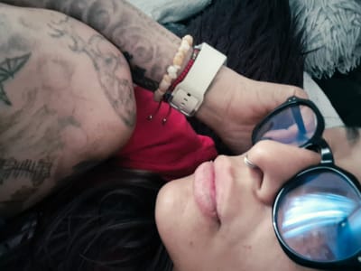 The image shows a close-up of a person lying down, with their face partially visible. They are wearing round, dark-rimmed glasses with blue lenses. A silver ring is on their nose. Their lips are full and a light pink color. To the left, their arm is visible, covered in tattoos. They are also wearing a white watch and two bracelets, one with light-colored beads and another with small red beads. A bright red garment is visible beneath their arm. The background is dark and appears to be fabric, possibly a blanket or clothing. The overall impression is of a relaxed, personal moment. No specific location in Bridlington, United Kingdom can be determined from this image.