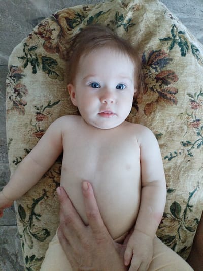A baby with blue eyes and light brown hair lies on a patterned fabric. The baby is wearing a diaper and is mostly unclothed. A hand is visible on the baby's torso. The setting appears to be indoors, with a textured floor visible around the fabric. The fabric has a floral pattern in muted colors. The baby's expression is neutral. There is no visible text in the image. The location context is Vinogradny, Russia.