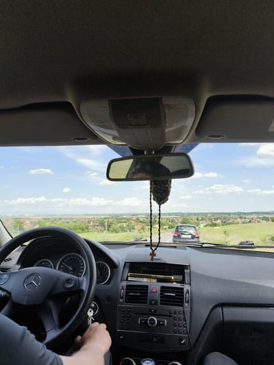 The image captures the view from inside a moving Mercedes-Benz car, likely a C-Class model. The driver's hands are visible on the steering wheel, with keys in the ignition. The dashboard features a classic Mercedes design with analog gauges and a central control panel. A rosary with a cross hangs from the rearview mirror, along with a skull-shaped ornament. Through the windshield, the scene reveals a straight road with another car ahead. The surrounding landscape features a blend of residential areas with red-tiled roofs and green fields under a partly cloudy sky, indicative of a rural or suburban setting. Based on the information provided, the location is likely Isperihovo, Bulgaria.