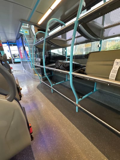 The image depicts the interior of a public transport bus, likely an intercity or airport coach, specifically located in Crumlin, United Kingdom. The perspective is from a passenger seat in the mid-section, looking forward and slightly right towards the luggage area and the front of the vehicle.
In the foreground, the back of a light grey passenger seat is partially visible on the left. The bus aisle, featuring a two-tone speckled floor (lighter grey-brown on the left, darker grey on the right), extends towards the front. To the right of the aisle, a dedicated luggage storage area runs along the side of the bus. This area consists of two tiers of racks framed with light blue/teal colored metal tubing and featuring perforated mesh shelving.
On the lower tier of the luggage rack, an olive-green hardshell suitcase is positioned horizontally, with a white paper tag bearing the text "BFS RC191" and a barcode attached to its right side. Next to it, to the left, a black soft-sided bag or backpack rests. Further down the aisle, a small black hardshell suitcase is visible on the lower rack.
Through the bus windows on the right, an exterior urban environment can be seen, including parts of buildings with lighter facades. Overhead, the ceiling features recessed fluorescent lighting strips. Towards the front of the bus, a set of steps with light blue/teal handrails leads to what appears to be an elevated section or the driver's cabin area. The driver's area includes blue and grey panels and electronic displays. No individuals are present in the visible scene.