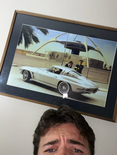 The image displays a large, framed color photograph mounted at an angle on a plain, light-colored wall. The frame consists of a dark blue matboard and a thin, brown-toned wooden border. The photograph depicts a vintage silver Chevrolet Corvette Sting Ray coupe, likely a 1960s model, parked outdoors on an asphalt surface. In the background of the photograph stands the iconic Theme Building at Los Angeles International Airport (LAX), characterized by its distinctive parabolic arches and central saucer-shaped observation deck. Two individuals are visible in the photograph: a male dressed in a pilot's uniform, complete with a cap, and a female in what appears to be a flight attendant's uniform, both standing near the rear of the vehicle. Palm trees are also present within the background of the framed image, suggesting a warm climate.
Below the framed photograph, occupying the bottom portion of the overall image, are the upper facial features of an individual. Only the eyes, eyebrows, and forehead are visible. The individual's eyes appear moist, and their brow is slightly furrowed. The hair is dark and styled with some volume.
The physical location of the wall and the framed photograph within the city of Rancho Palos Verdes is not ascertainable from the provided visual information. The location depicted within the framed photograph is Los Angeles International Airport, Los Angeles, United States.