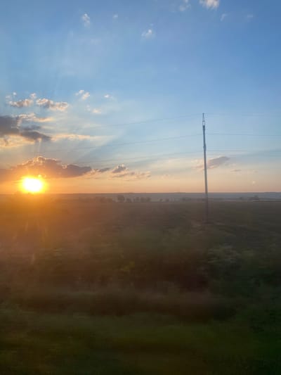 The image captures a sunset over a rural landscape, likely near Balabanu, Moldova. The sun, a bright orb, is positioned low on the horizon, casting golden rays through scattered clouds. The sky is a gradient of blue and orange, transitioning to a soft pinkish hue. In the foreground, a dark, textured field stretches out, appearing to be a cultivated crop. A tall utility pole with power lines is a prominent vertical element on the right side of the frame, silhouetted against the colorful sky. In the distance, a hazy line of trees and structures suggests a small settlement or agricultural buildings. The overall impression is one of natural beauty and a serene rural evening.