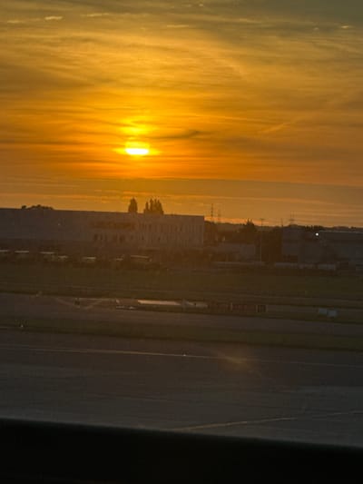 The image shows a sunset over a landscape that is likely near Brussels Airport in Zaventem, Belgium. The sky is filled with hues of orange and yellow from the setting sun, with wispy clouds scattered across it. In the midground, a long, flat-roofed building with numerous windows is visible, possibly a cargo facility or an airport terminal building. A row of vehicles, some appearing to be trucks or buses, are parked in front of this building. Further in the distance, there are hints of industrial structures and possibly utility poles. The foreground is dominated by what appears to be a paved surface, possibly a runway or tarmac, with some faint markings. The overall impression is one of an industrial or airport environment at dusk, with the warm glow of the sunset illuminating the scene. There are no people clearly visible in the image.