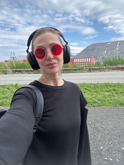 This media file captures a person wearing headphones and red-tinted sunglasses, taking a selfie outdoors. The person is dressed in a black long-sleeved shirt and has a backpack on. Their face is partially visible, with a nose ring and a tattoo on their neck.
The setting appears to be in Norilsk, Russia, suggested by the industrial buildings and the mountainous, sparsely vegetated landscape in the background. The sky is partly cloudy, indicating it could be daytime. The ground is paved, suggesting a road or pathway, with grass growing along the side. The overall scene conveys a sense of outdoor activity, possibly walking or exploring in a somewhat industrial or remote environment. There is no visible text in the image.