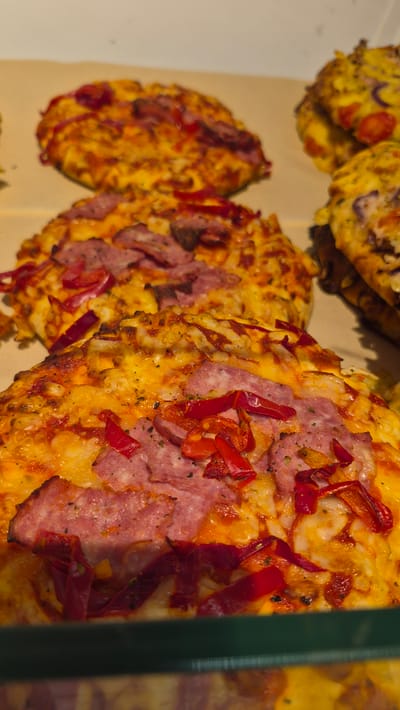 The image displays a close-up view of several small, round pizzas arranged in a line. The pizzas have a golden-brown crust, generously covered with melted cheese. Visible toppings include slices of cured meat, likely salami, and strips of red bell pepper. Some scattered dark specks, possibly herbs or spices, are also present. The pizzas appear to be resting on a light-colored surface, possibly parchment paper or a similar baking material. The lighting suggests an indoor setting, likely a bakery or food establishment, with a green vertical element on the right edge of the frame, which could be part of a display case or shelf. The focus is sharp on the pizzas in the foreground, with the ones further back becoming progressively blurred. There is no visible text in the image. The pizzas seem to be freshly prepared or ready for sale. The scene evokes a sense of appetite appeal due to the rich colors and textures of the food.