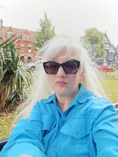 A medium shot selfie features a woman with long, very light blonde hair and prominent sunglasses with tortoiseshell frames. She is wearing a bright blue button-down shirt with two chest pockets. Her lips are painted a light pink. The background shows a park-like setting with lush green foliage in the foreground, including a large palm-like plant. In the mid-ground, there are several buildings: on the left, a red brick building with arched windows and gables, and on the right, a Tudor-style building with dark timber framing and white infill. Two cars are visible on a street behind the park, one silver and one red. The sky is overcast and bright, suggesting daytime with perhaps cloudy weather. The overall impression is a casual outdoor portrait in a semi-urban environment, possibly taken during pleasant weather. The location is specified as Bedford, United Kingdom. There is no visible text in the image.