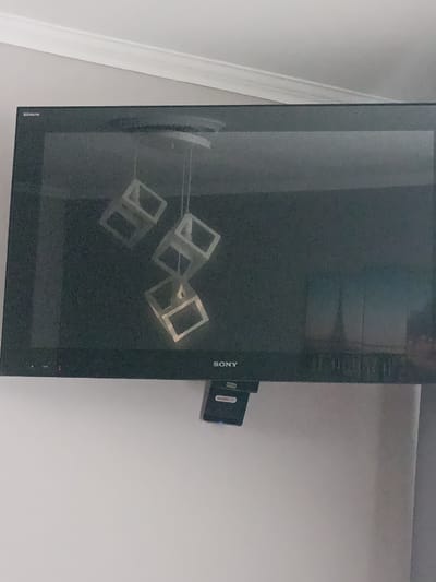 The image shows a television mounted on a light gray wall. The television screen displays a reflection of four hollow cubes suspended in mid-air, appearing to be part of a mobile. The cubes are a light color and have open sides. Below the television, a small black device with some text is attached. The top of the image shows white molding along the wall. The overall scene appears to be an indoor setting, likely a room in a home. The lighting suggests it is daytime, but no specific time can be determined. The image is grainy, which obscures some finer details. Visible text on the television reads "BRAVIA" and "SONY". The small black device below the TV has text that appears to read "Snell TV".